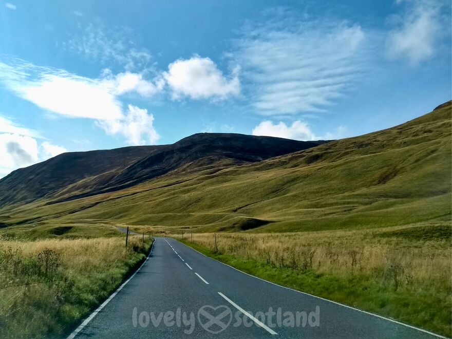 Scenic Highland road through rolling green hills under a bright blue sky