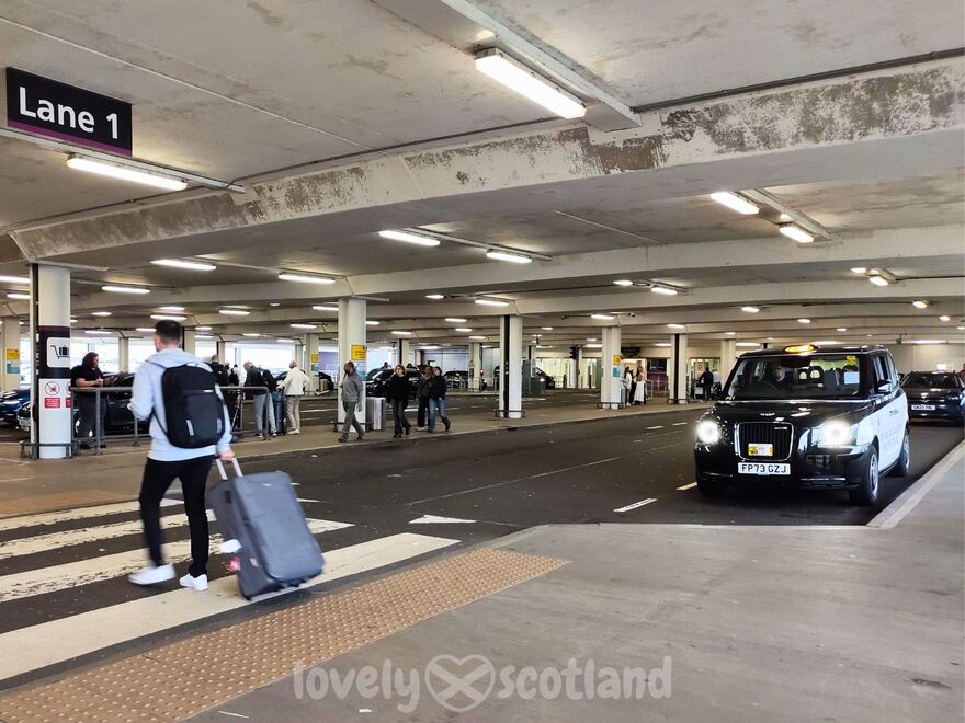 Black taxi at Edinburgh Airport rank beside a traveler crossing with a suitcase